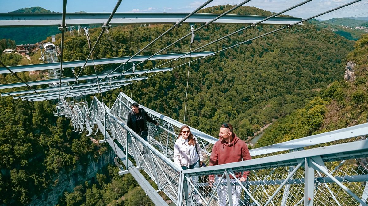 Посетители идут по подвесному мосту Skybridge над зелёным ущельем в ясный день